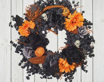 May include: A circular wreath featuring black foliage, vibrant orange and black fabric flowers, and small white skull accents. The arrangement includes orange glittered fern-like stems, black glittered leaves, and textured orange spheres integrated throughout the dark base.