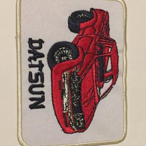 May include: Red Datsun pickup truck embroidered patch with white background and a cream colored border.