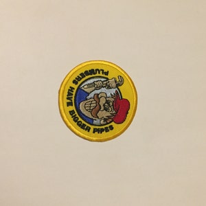May include: A yellow embroidered patch with a cartoon plumber holding a wrench. The text on the patch reads "Plumbers Have Bigger Pipes".