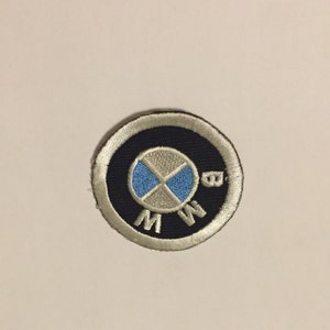 May include: A round, white and blue embroidered patch with the BMW logo. The logo is a white circle with a blue center and a white propeller design. The words "BMW" are embroidered in white around the circle.