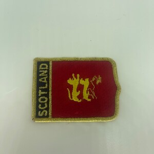 Op de afbeelding: Een geborduurde patch in rood en goud met het woord "SCOTLAND" en een leeuw in het wapen.