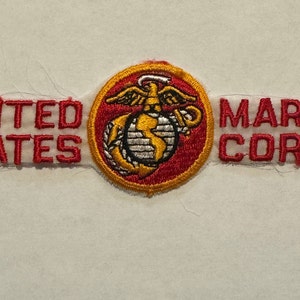 Könnte beinhalten: Gestickter Aufnäher mit dem Text "UNITED STATES MARINE CORPS" in roter Schrift. Das zentrale Emblem ist ein goldener und weißer Globus mit Adler und Anker, vor einem roten und gelben kreisförmigen Hintergrund.