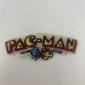 以下が含まれることがあります： 黄色、赤、黒のステッチで刺繍されたビンテージのパックマンのワッペン。ワッペンには、象徴的なパックマンのキャラクターと、ミッドウェイとCBSのロゴが付いた「PAC-MAN」の文字が描かれています。