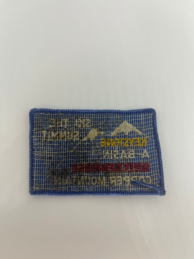 May include: A rectangular blue patch with white text and a mountain graphic. The patch reads "Ski the Summit" and "Keystone" in yellow and red lettering. The patch is bordered with blue stitching.