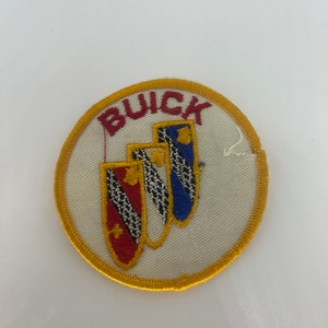 Peut inclure: Écusson brodé avec le mot "BUICK" en rouge au-dessus de trois écussons. Les écussons sont rouges, blancs et bleus avec des détails dorés, sur un fond crème avec une bordure dorée. L'écusson est circulaire.