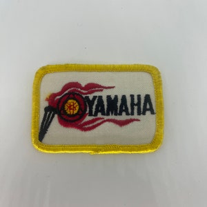 May include: A yellow embroidered patch with a red and black Yamaha logo. The logo features a stylized flame with a circle containing a tuning fork inside.