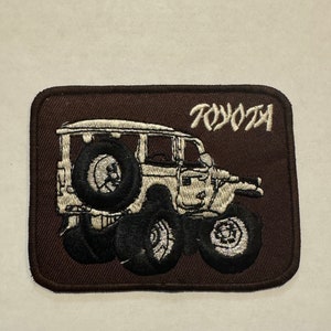 Könnte beinhalten: Ein braunes gesticktes Patch mit einem weißen Toyota Land Cruiser und dem Schriftzug "TOYOTA" in weißen Buchstaben.