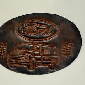 May include: Oval, brown leather belt buckle featuring a Ford van design. The Ford logo is embossed at the top, with a van below and leaf designs on either side. The buckle has a dark brown border.