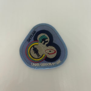 May include: A light blue Skylab patch with embroidered details. The patch features a circular design with a silhouette of a person, a stylized planet, and the text "SKYLAB" and "CARR GIBSON POGUE". The patch has a blue border.