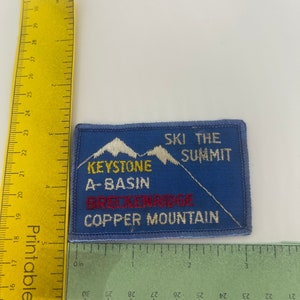 May include: Blue rectangular patch with white embroidered text and mountain graphics. The patch reads "SKI THE SUMMIT" and lists "KEYSTONE A-BASIN BRECKENRIDGE COPPER MOUNTAIN". A yellow ruler is placed next to the patch.