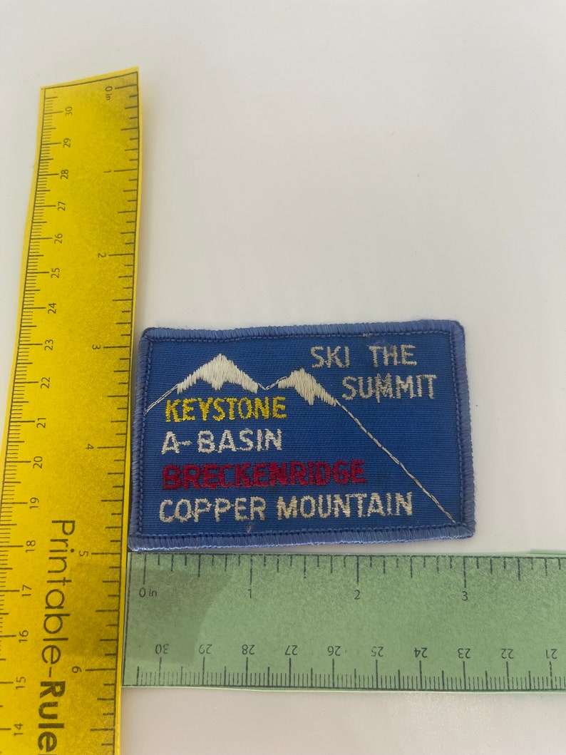 May include: Blue rectangular patch with white embroidered text and mountain graphics. The patch reads "SKI THE SUMMIT" and lists "KEYSTONE A-BASIN BRECKENRIDGE COPPER MOUNTAIN". A yellow ruler is placed next to the patch.