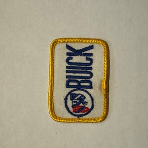 Peut inclure: Un écusson en tissu blanc avec une bordure jaune. L'écusson porte le mot "BUICK" en lettres bleues et un logo bleu et rouge au centre.