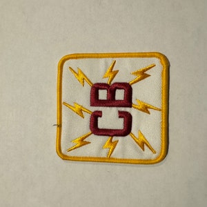Könnte beinhalten: Gestickter Aufnäher mit weißem Hintergrund, goldenem Rand und den Buchstaben "CB" in Burgunderrot. Gelbe Blitze strahlen von hinter den Buchstaben aus. Der Aufnäher ist quadratisch mit abgerundeten Ecken.