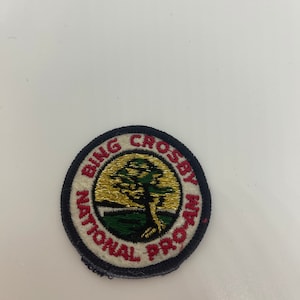 May include: A round, white patch with black stitching. The patch has a red border and the words "Bing Crosby National Program" in red lettering. A gold and green tree is in the center of the patch.