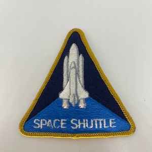 May include: Embroidered patch with a white space shuttle design on a navy blue and light blue triangular background. The words "SPACE SHUTTLE" are embroidered in white. The patch has a gold border.