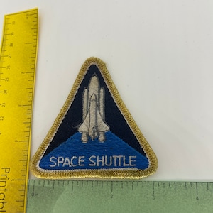 May include: An embroidered patch in the shape of a triangle with a gold border. The patch depicts a white space shuttle against a blue background with the text "SPACE SHUTTLE" in gold lettering.