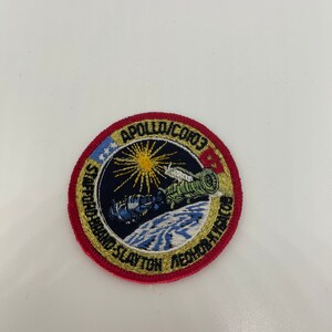 Peut inclure: Un écusson brodé rouge, blanc et bleu avec le texte "APOLLO-SOYUZ TEST PROJECT" et "DONALD SLAYTON - ALEXEY LEONOV" autour du bord. Le centre de l'écusson représente un vaisseau spatial en orbite autour de la Terre.