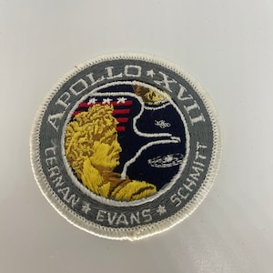 Może przedstawiać: Naszywka haftowana w kolorze szarym i białym z tekstem "Apollo XVII" i nazwiskami "Cernan, Evans, Schmitt". Naszywka przedstawia postać astronauty w kolorze żółtym i brązowym na niebieskim i białym tle.