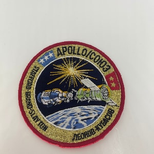 May include: A gold and red embroidered patch with the text "Apollo-Soyuz Test Project" and "Apollo-Soyuz Test Project" in a circle around a depiction of a spacecraft in orbit around Earth. The text "Apollo-Soyuz" is in a gold font and the text "Test Project" is in a black font. The patch also features a depiction of the American and Soviet flags.
