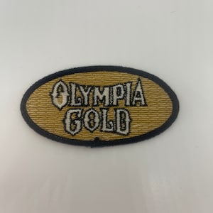 May include: A gold oval-shaped patch with black stitching around the edges. The words "OLYMPIA GOLD" are embroidered in white on the patch.