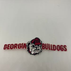 May include: A white and red embroidered patch with the words "GEORGIA BULLDOGS" and a cartoon bulldog wearing a red hat with a "G" on it.
