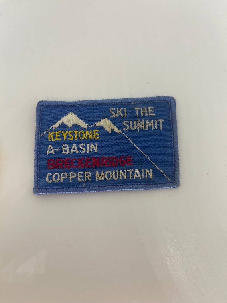 May include: A blue rectangular patch with white embroidered mountains and the text "SKI THE SUMMIT". Below the mountains are the words "KEYSTONE A-BASIN BRECKENRIDGE COPPER MOUNTAIN" in yellow and red lettering.