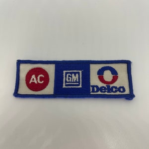 Może przedstawiać: Naszywka haftowana w kolorze białym, niebieskim i czerwonym z napisem "AC Delco" i logo General Motors.
