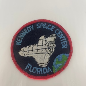 Pode incluir: Um remendo redondo com borda vermelha e fundo azul. O remendo apresenta um ônibus espacial branco, um globo terrestre azul e o texto "Kennedy Space Center Florida".