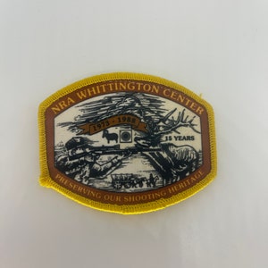 May include: A brown and yellow embroidered patch with the text "NRA Whittington Center" and "Preserving Our Shooting Heritage". The patch also features a black and white illustration of a person shooting a rifle with a deer in the background. The text "1973 - 1988" and "15 Years" are also included on the patch.