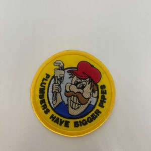 May include: A yellow embroidered patch with a cartoon plumber holding a wrench. The text around the plumber reads "Plumbers Have Bigger Pipes".