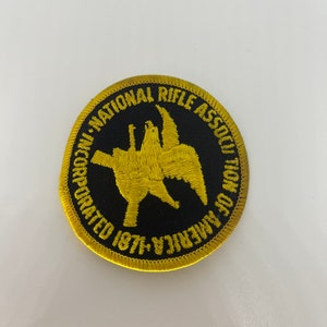May include: A circular patch with a black background and gold embroidered details. The patch features the text "NATIONAL RIFLE ASSOCIATION OF AMERICA" and a gold emblem. The text "INCORPORATED 1871" is also present.
