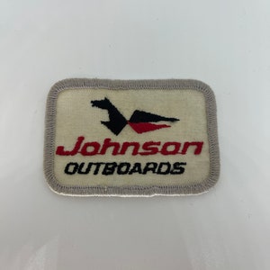 Puede incluir: Un parche de tela blanco con un borde gris. El parche presenta un logotipo de pájaro estilizado en negro y rojo y el texto "Johnson Outboards".