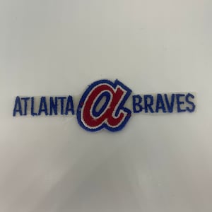 Puede incluir: Un parche blanco con las palabras "Atlanta Braves" en letras azules y rojas. Una "A" roja con un contorno azul está en el centro.