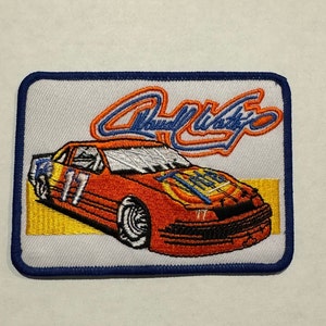 Könnte beinhalten: Ein gestickter Aufnäher mit einem roten Rennwagen mit der Nummer 17 an der Seite. Das Auto befindet sich auf einem gelben Hintergrund mit dem Namen "Rusty Wallace" in blauen und orangen Buchstaben.