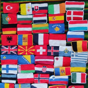 Könnte beinhalten: Eine Sammlung gehäkelter Flaggen aus verschiedenen Ländern, jede mit unterschiedlichen Farben und Designs. Die rechteckigen Flaggen sind eng angeordnet und zeigen eine Vielzahl von Nationalsymbolen und -mustern. Die Flaggen werden auf einer gestreiften grün, gelb und braunen Oberfläche präsentiert.