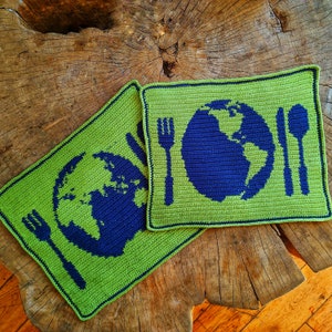 May include: Two green crocheted placemats with blue embroidered images of the Earth and a fork and knife. The placemats are on a wooden surface.