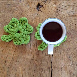 May include: Two green crocheted four-leaf clover coasters with a white ceramic mug filled with dark red liquid on top of one of the coasters.