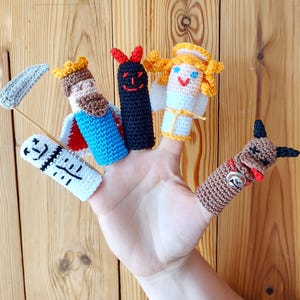 May include: A hand displaying six colorful, crocheted finger puppets. The puppets include a king, devil, angel, and a dog, each with unique features and expressions. The puppets are made of yarn in various colors, including blue, red, black, and brown.