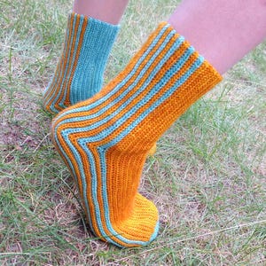 Puede incluir: Calcetines de punto con un diseño a rayas en naranja y azul claro. Los calcetines están hechos de un material texturizado. El diseño presenta bandas de colores alternas. Los calcetines se muestran sobre una superficie cubierta de hierba.