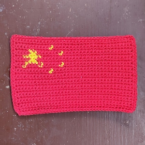 Op de afbeelding: Een handgemaakt, rechthoekig, rood gehaakt item met het ontwerp van de Chinese vlag. Het vlagontwerp bevat een grote gele ster en vier kleinere gele sterren in de linkerbovenhoek.