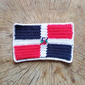 Puede incluir: Bandera de la República Dominicana hecha a crochet, con los colores rojo, blanco y azul, con un emblema verde en el centro.