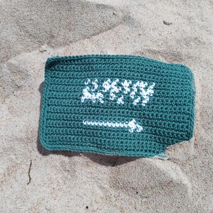 Puede incluir: Una bolsa rectangular de ganchillo verde azulado con texto blanco. La bolsa está sobre una superficie arenosa. El texto blanco parece ser una palabra o frase estilizada.