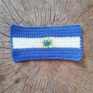 May include: A crocheted blue and white rectangular item with a central emblem. The item features a blue border, a white horizontal stripe, and a central emblem with green and yellow details. The item is placed on a wooden surface.