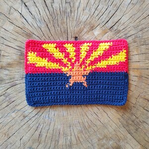 May include: Crocheted Arizona state flag with a red, yellow, and orange sun design on a blue background.
