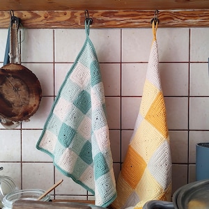 May include: Two handmade crochet kitchen towels hanging on hooks. One towel features a patchwork design in shades of teal, white, and pink. The other towel has a yellow and white patchwork design. The towels are hanging against a tiled wall.