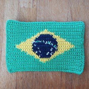 以下が含まれることがあります： ブラジル国旗をモチーフにしたかぎ針編みの長方形のアイテム。デザインは、緑色の背景、黄色のひし形、白い星と湾曲したバナーが付いた青い円で構成されています。このアイテムはおそらく装飾品です。