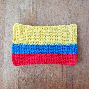May include: A rectangular, handmade crochet item featuring horizontal stripes of yellow, blue, and red. The item is placed on a wooden surface, showcasing the vibrant colors and textured stitches of the crochet work.