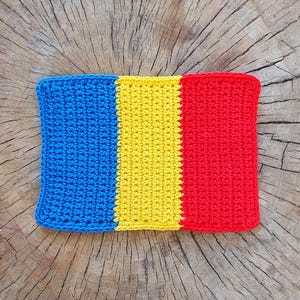 May include: A crocheted flag of Romania, with blue, yellow, and red stripes, on a wooden background.