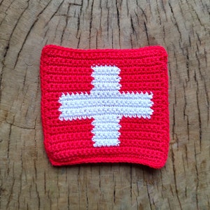 May include: A crocheted square with a white cross on a red background. The square is made of yarn and has a textured surface.