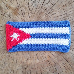 May include: A crocheted rectangular item featuring the Cuban flag design. The item has a blue border, white and blue stripes, and a red triangle with a white star. The item is placed on a wooden surface.